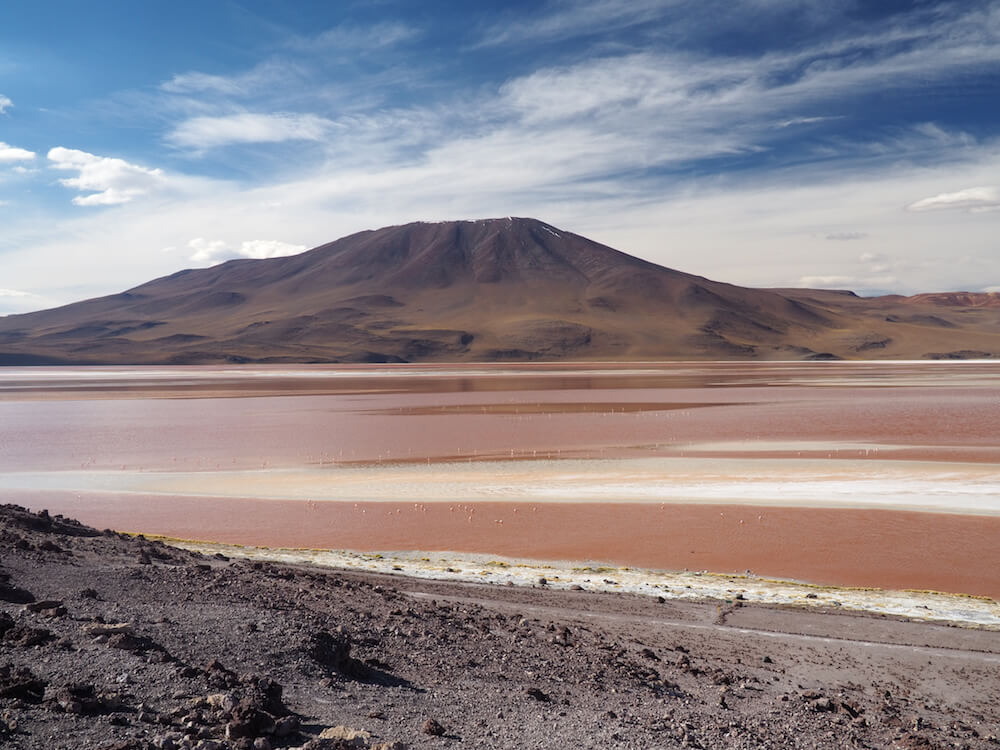 A pink lake sits between rocks in the foreground and a volcano in the background