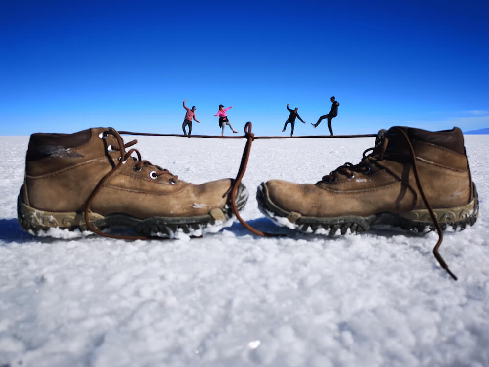 Two brown walking boots placed on the salt flat with four people seemingly "balancing" on the laces like a tightrope