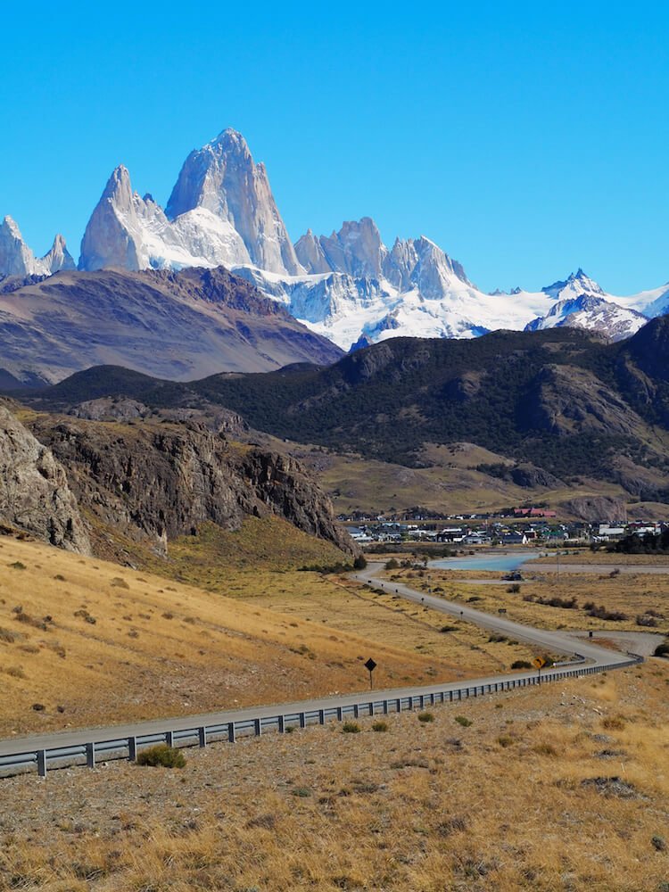 A winding road leads to a small town surrounded by craggy and snow-covered mountains