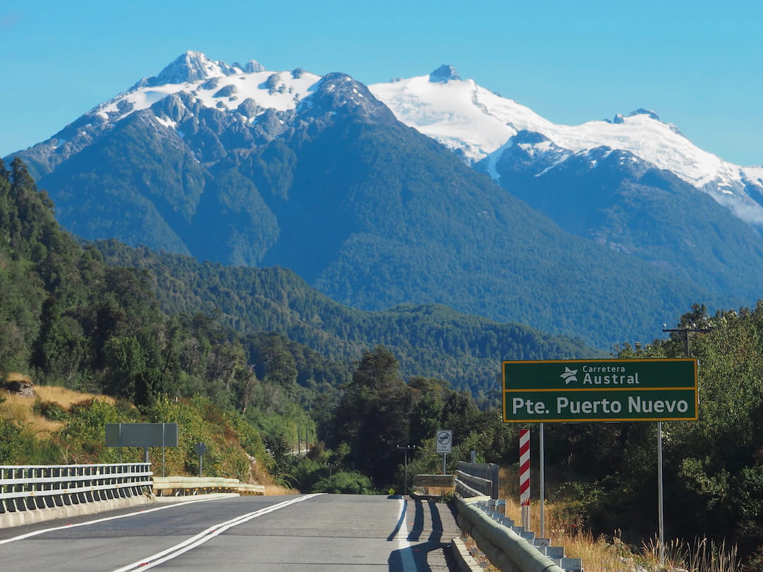 A green road sign stands beside a tarmac road with trees and a snow-capped mountain in the background
