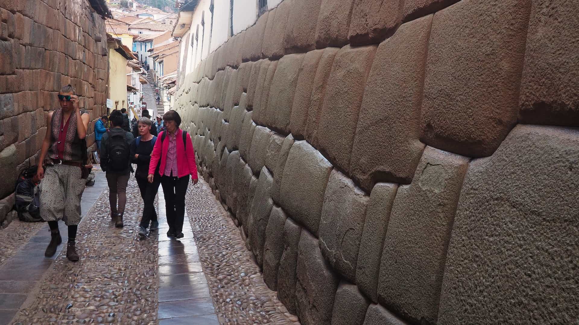 People walking in a narrow street with a large stone wall on the right