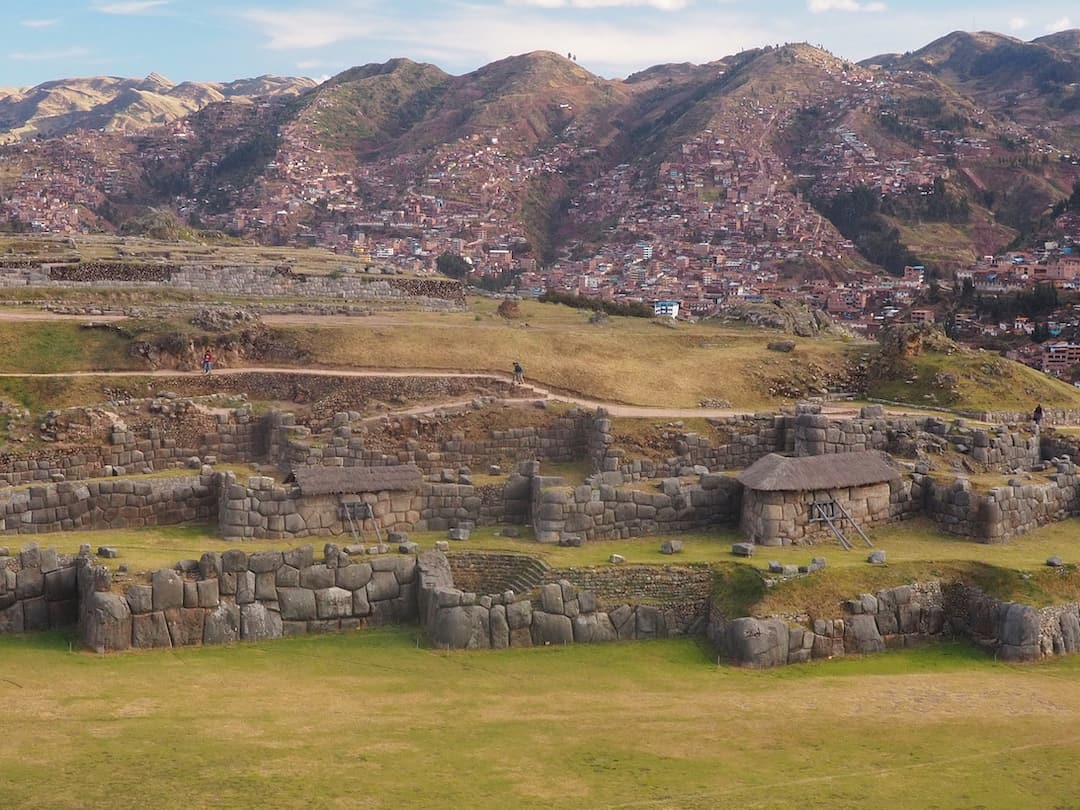 A panoramic view of ruined stone walls with red roofed houses and mountains in the background