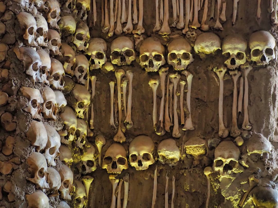 Human skulls on a wall facing the camera in yellow light and an adjacent wall facing side-on in natural light