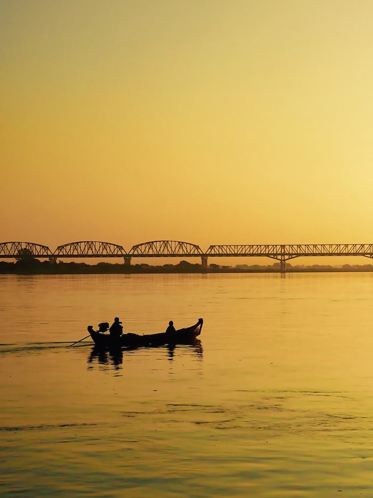 A small boat and a bridge are silhouetted against a yellow background of the sky and river