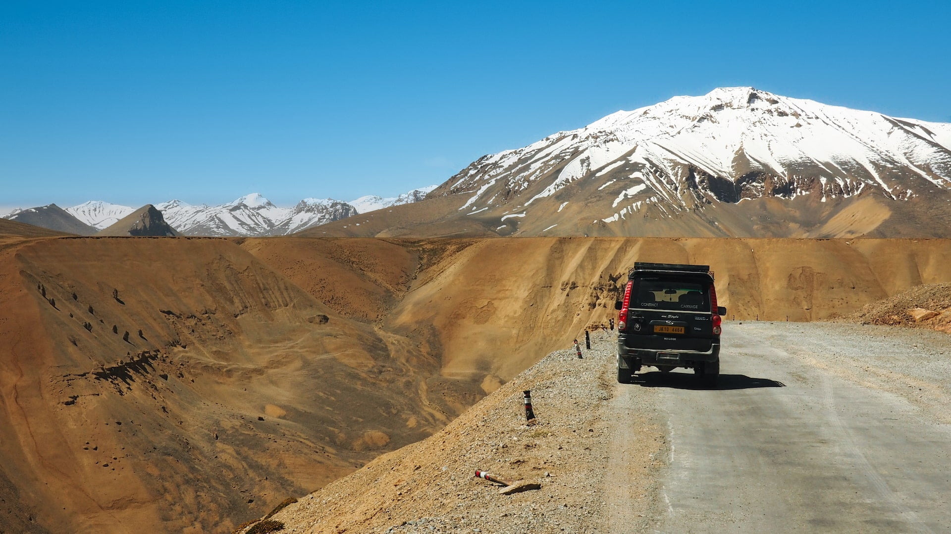 A car is parked on the side of a mountain road with a snow-capped peak in the background