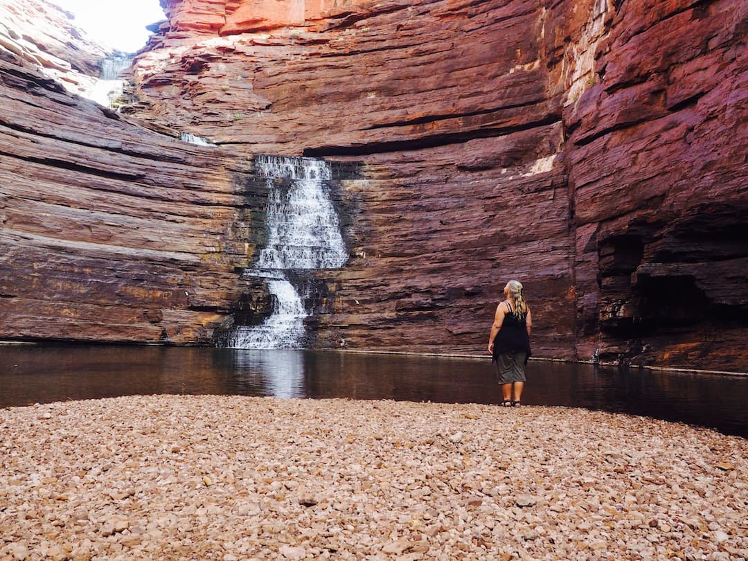 Nicky stands on a gravel beach looking upward to a waterfall