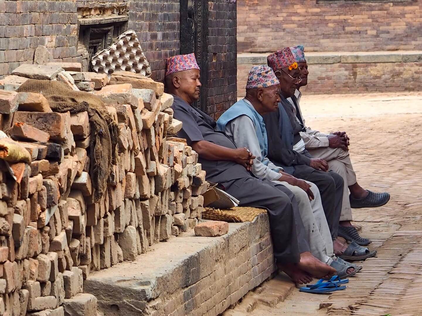 A pile of bricks in the foreground lead to four men sitting at the side of a street