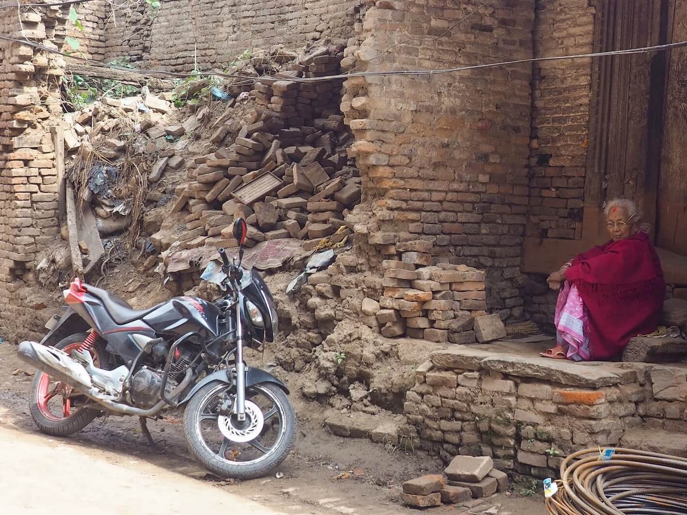 A woman in red clothing sits in a doorstep next to rubble and a motorcycle