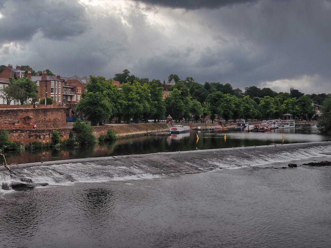 A river in the foreground with sandstone buildings, a wall and trees in the background