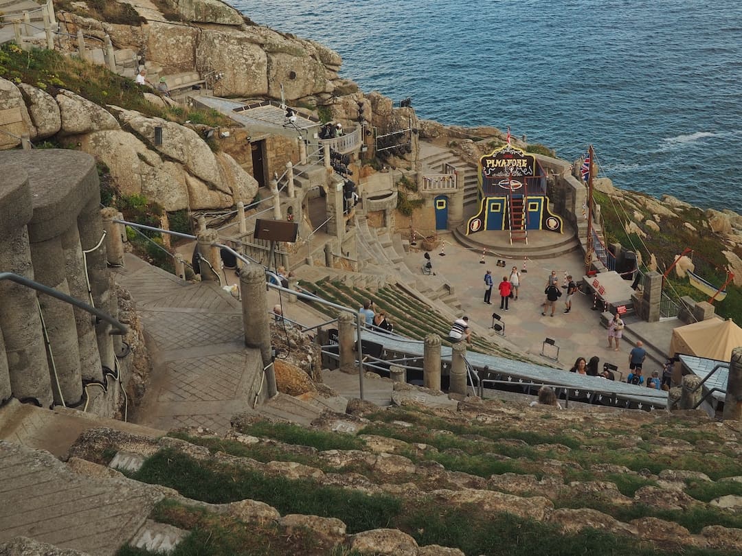 A view looking down a cliffside into a stone-seated theatre with the sea in the background