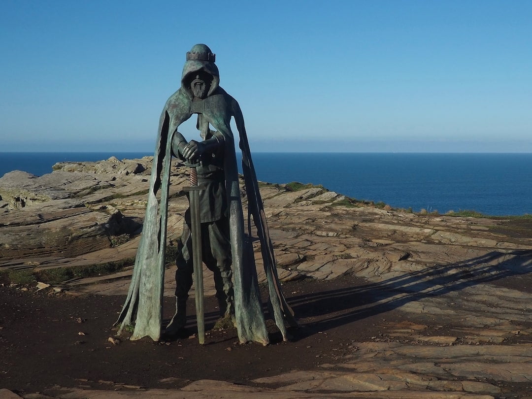 A metal statue of a king holding a sword stands on a cliff over looking the sea in the background