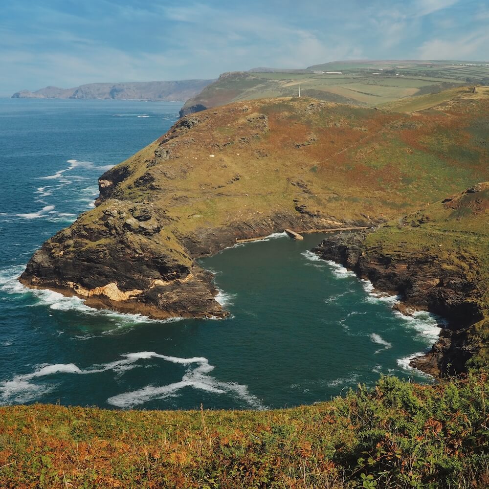A green-covered headland wraps around blue water of the sea, with hills in the background
