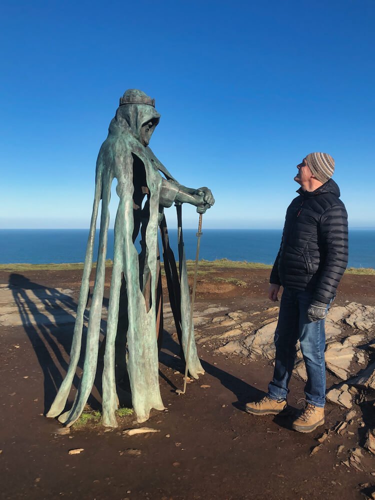 A statue of a man holding a sword on a clifftop with the sea in the background + a man staring up at it