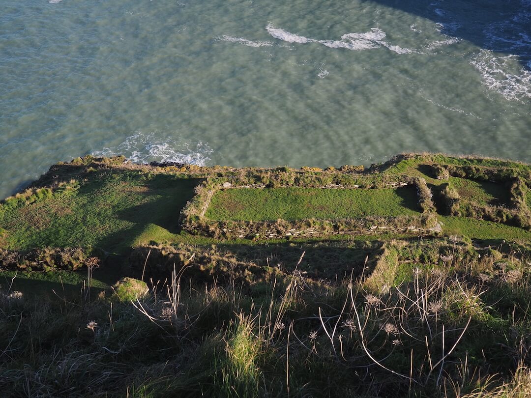 A patchwork of ancient building foundations and green grass sit sit on a hillside overlooking the sea