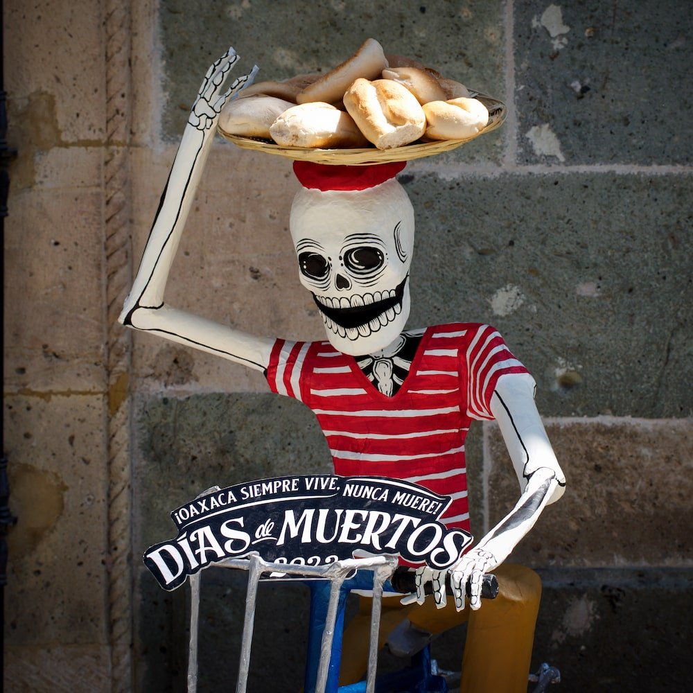 A skeleton in a red hooped shirt sits on a bicycle with a basket of bread on its head