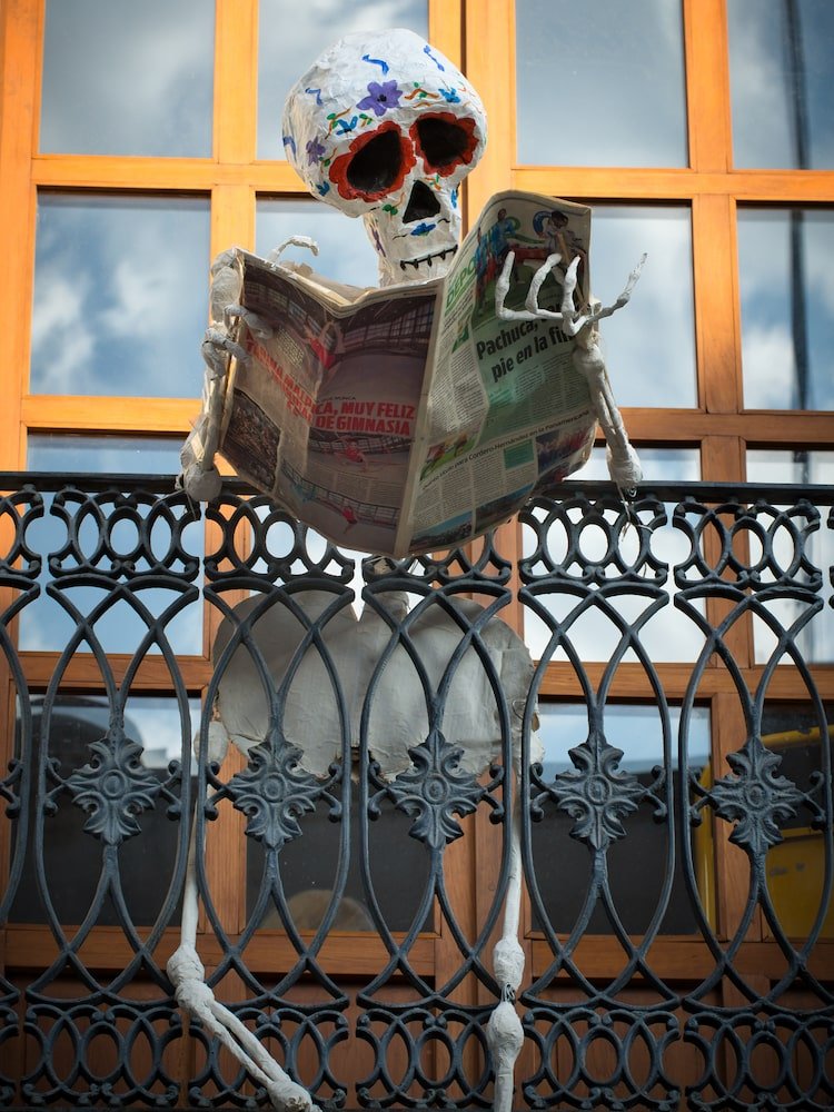 A skeleton stands on a balcony reading a newspaper