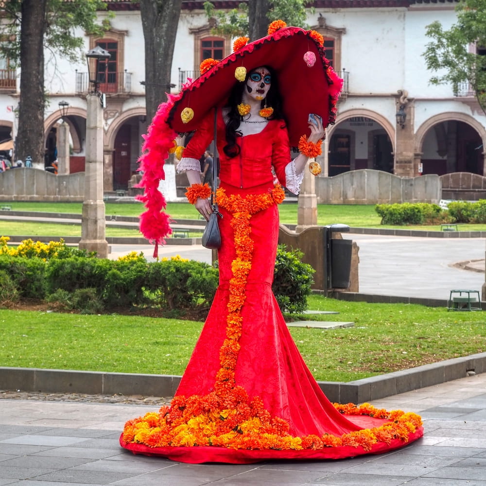 A catrina in a red dress poses in a plaza 